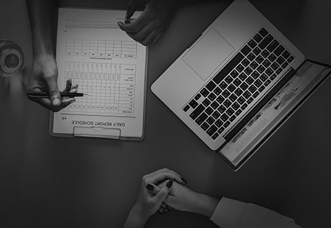 dos personas sentadas en una mesa, donde sólo se observa, desde la parte de arriba, sus manos, un bloc de notas y una laptop