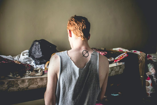 Muchacho pelirrojo de espaldas con un tatuaje en la nuca y una playera rota en una habitación desordenada y sucia