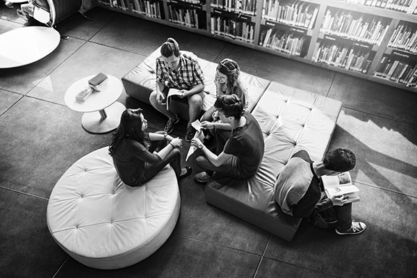 compañeros de escuela en una biblioteca platicando y un compañero apartado leyendo solo.