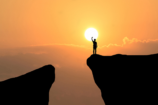 hombre en la cima de una montaña levantando el brazo derecho y de fondo la puesta del sol.